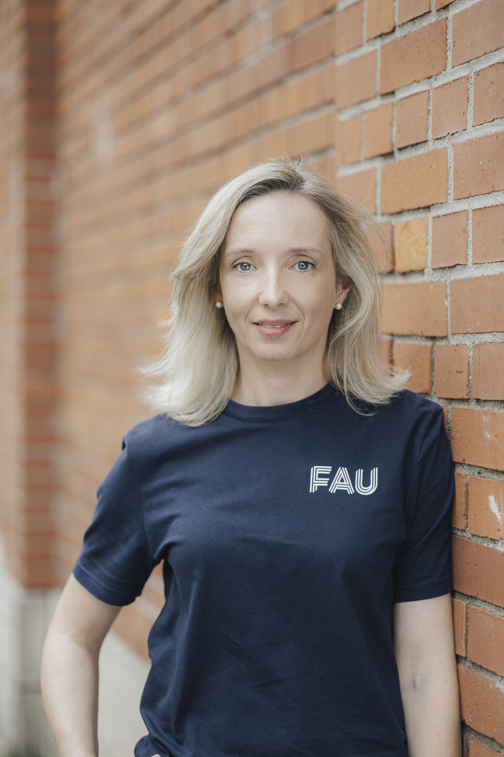A poirtrait picture of Prof. Kathrin Castiglione (female) in a marine blue tshirt with the FAU logo in front of the brick wall